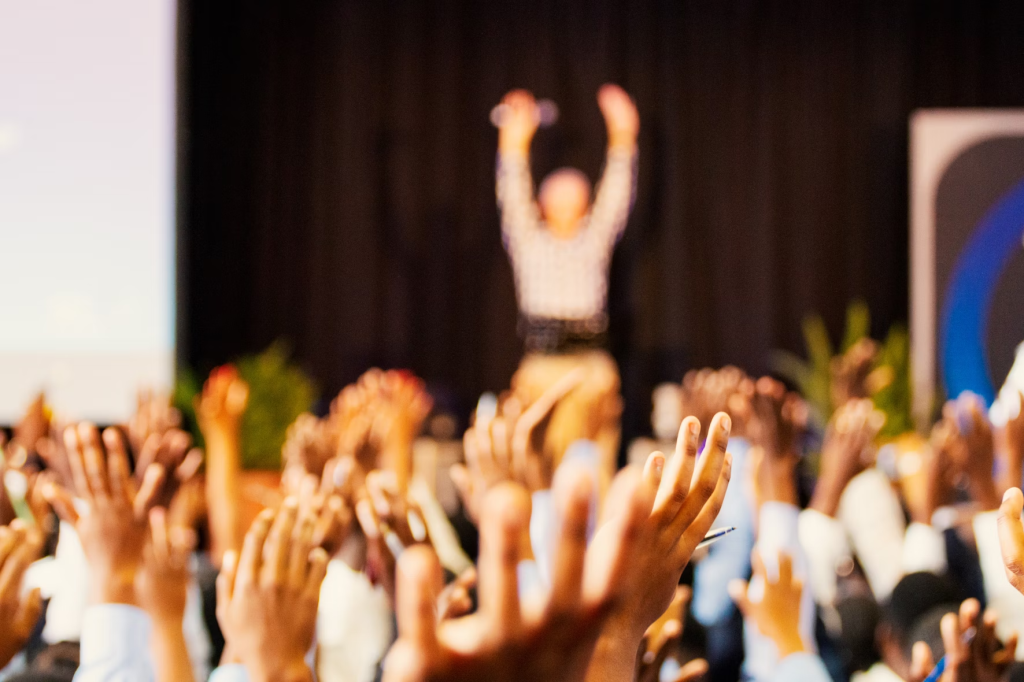 Conference room full of people with the focus on the audiences hands in the air and a blurry figure on a stage jumping with their hands in the air